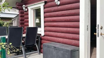 A log cabin with a patio area featuring two wicker tables and chairs, a small window with a white frame, and a door with a brass handle.