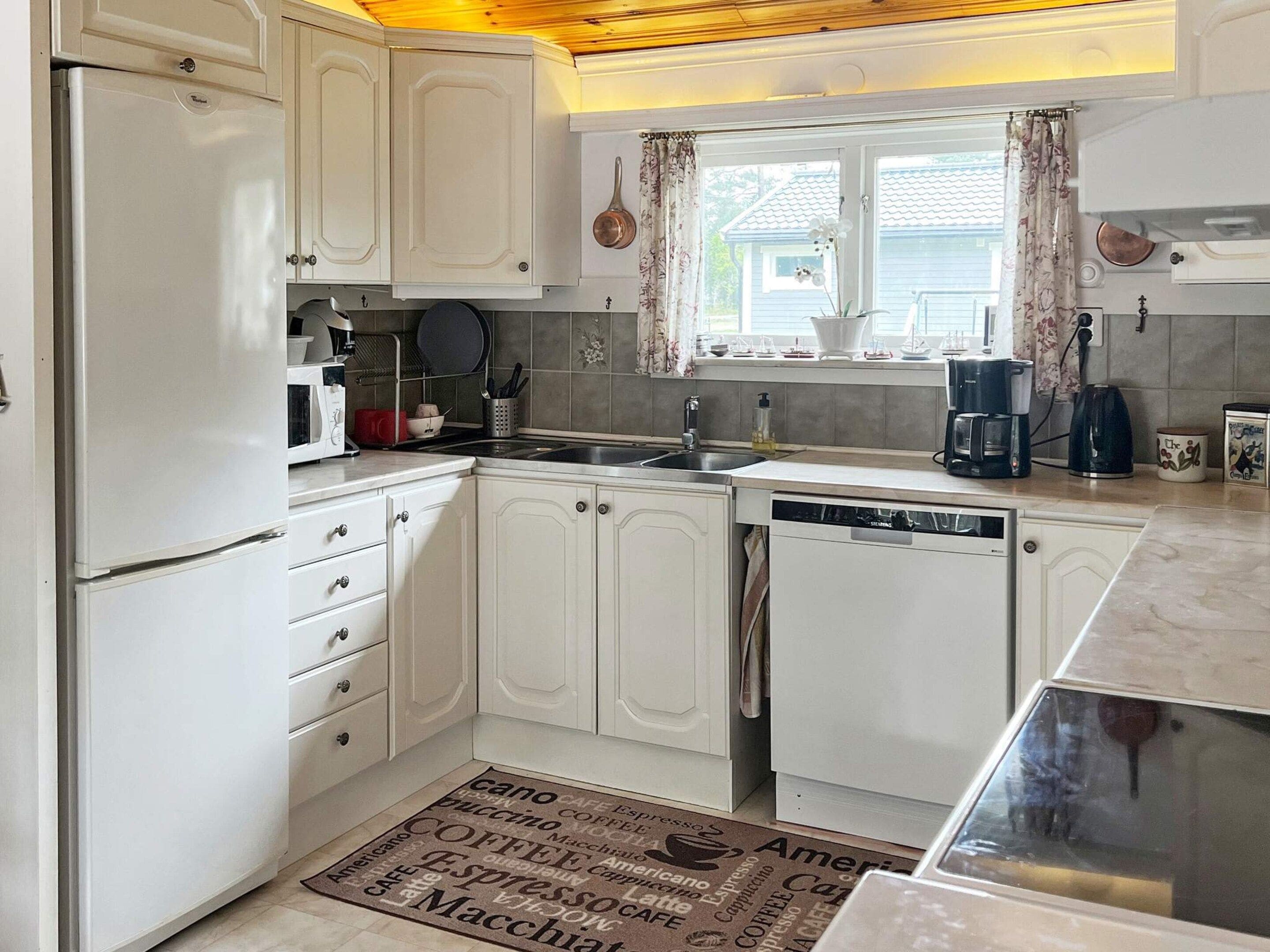 A compact kitchen with white cabinetry, a built-in dishwasher, and a window with curtains.