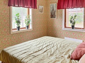 A bedroom with a bed, two windows with potted plants, floral wallpaper, and red curtains.