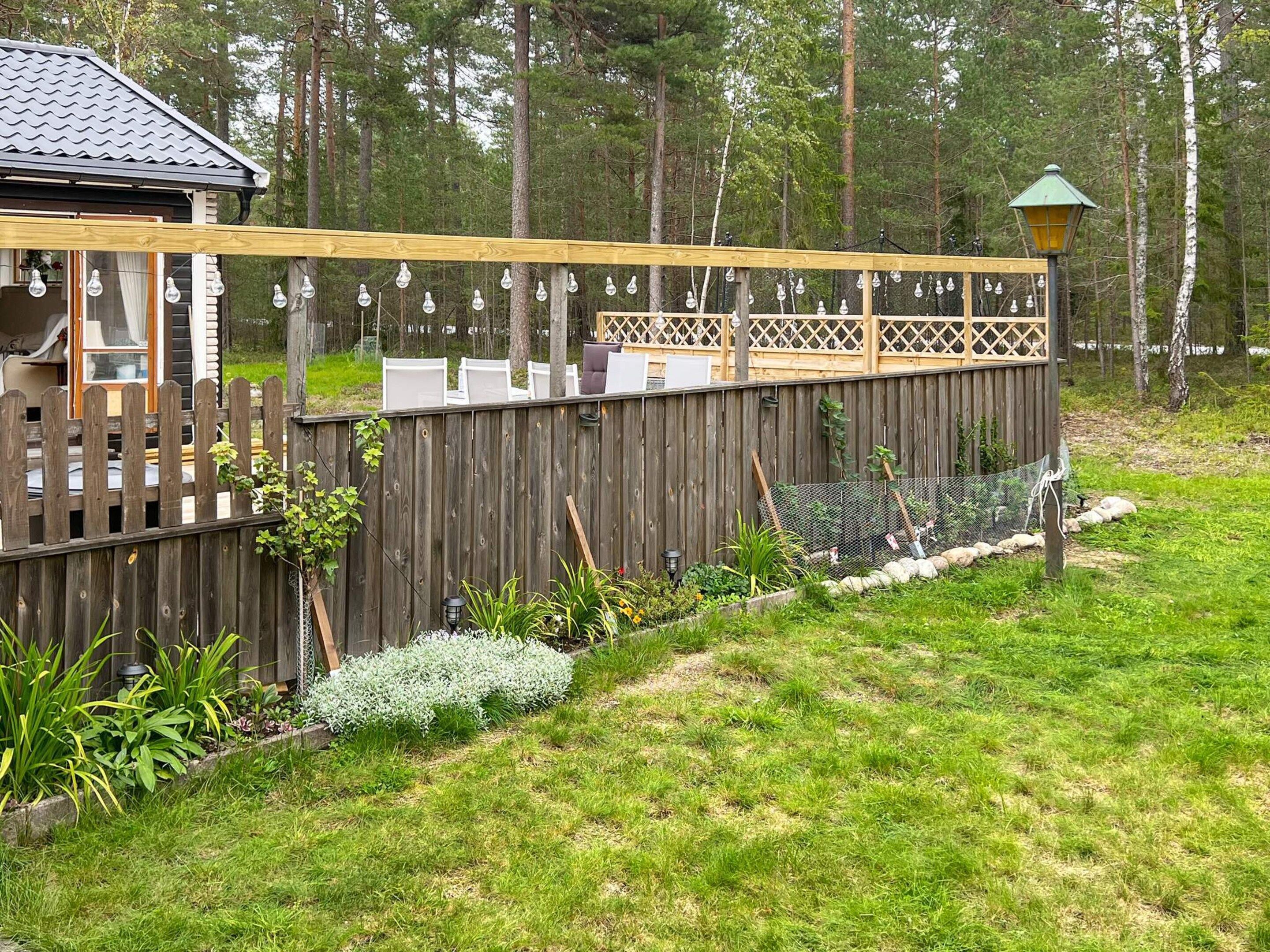 A wooden deck with a lattice railing, surrounded by a garden with various plants and a fence.