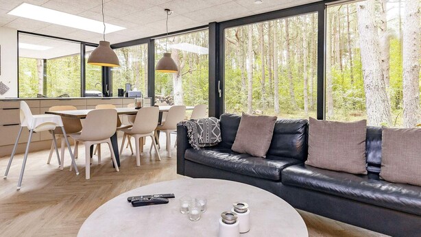 Una sala de estar moderna con un sofá de cuero negro, una mesa redonda de centro y un comedor con mesa y sillas. Amplias ventanas ofrecen vistas a un área boscosa.