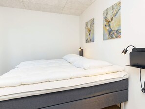 A neatly made bed with a grey mattress and white bedding in a room with white walls. There are two wall-mounted paintings and a black desk lamp on the right side.
