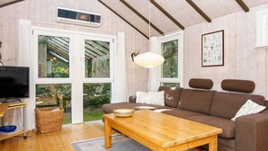 A cozy living room with a brown sofa, a wooden coffee table, and a television. There are large windows with curtains, a hanging light fixture, and a framed botanical print on the wall.