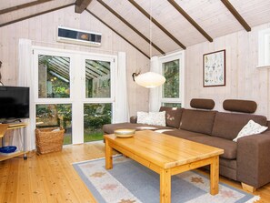 Ett mysigt vardagsrum med en brun soffa, ett träbord och en tv. Det finns stora fönster med gardiner, en hängande lampa och en inramad botanisk tavla på väggen.