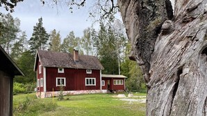 Une maison en bois rouge, avec un toit en pente et une cheminée, entourée d’une cour verdoyante et d’arbres.