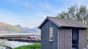 A small wooden cabin with a single window and a door, situated near a rocky shoreline with boats docked in the background.