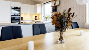 A modern kitchen with white cabinets, a central island, and a dining area with a vase of dried flowers. - 5 Star Holiday Home in Vaeggerlose (Vaeggerlose)