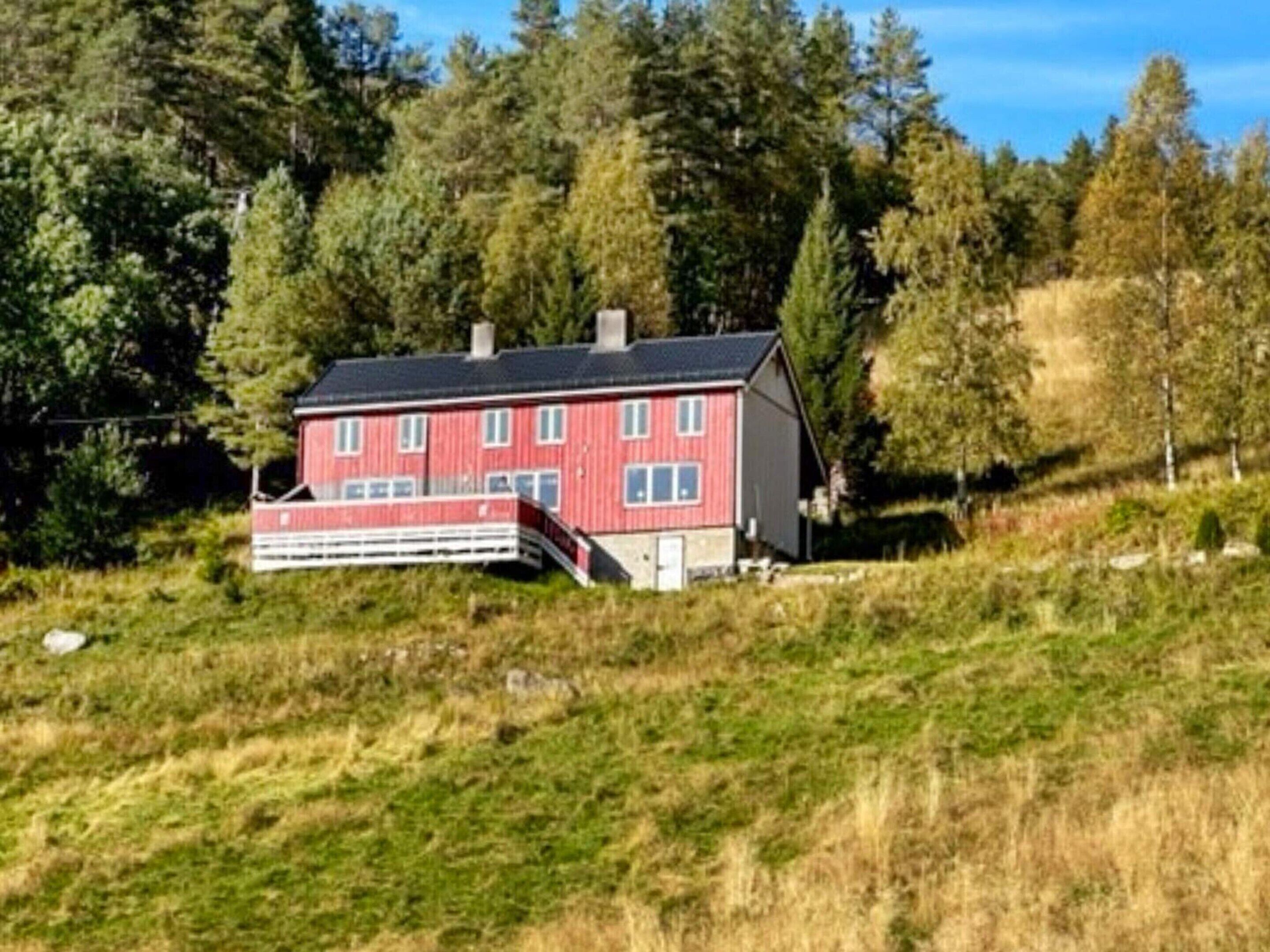 A red wooden house with a balcony, surrounded by trees and grassy terrain.
