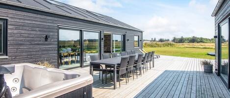 A modern outdoor deck with a hot tub, dining area, and large sliding glass doors offering a view of a grassy field.