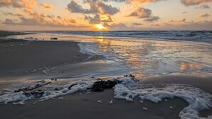 Escena de playa con el sol poniéndose sobre el océano, olas suaves lamiendo la orilla y rocas dispersas sobre la arena.