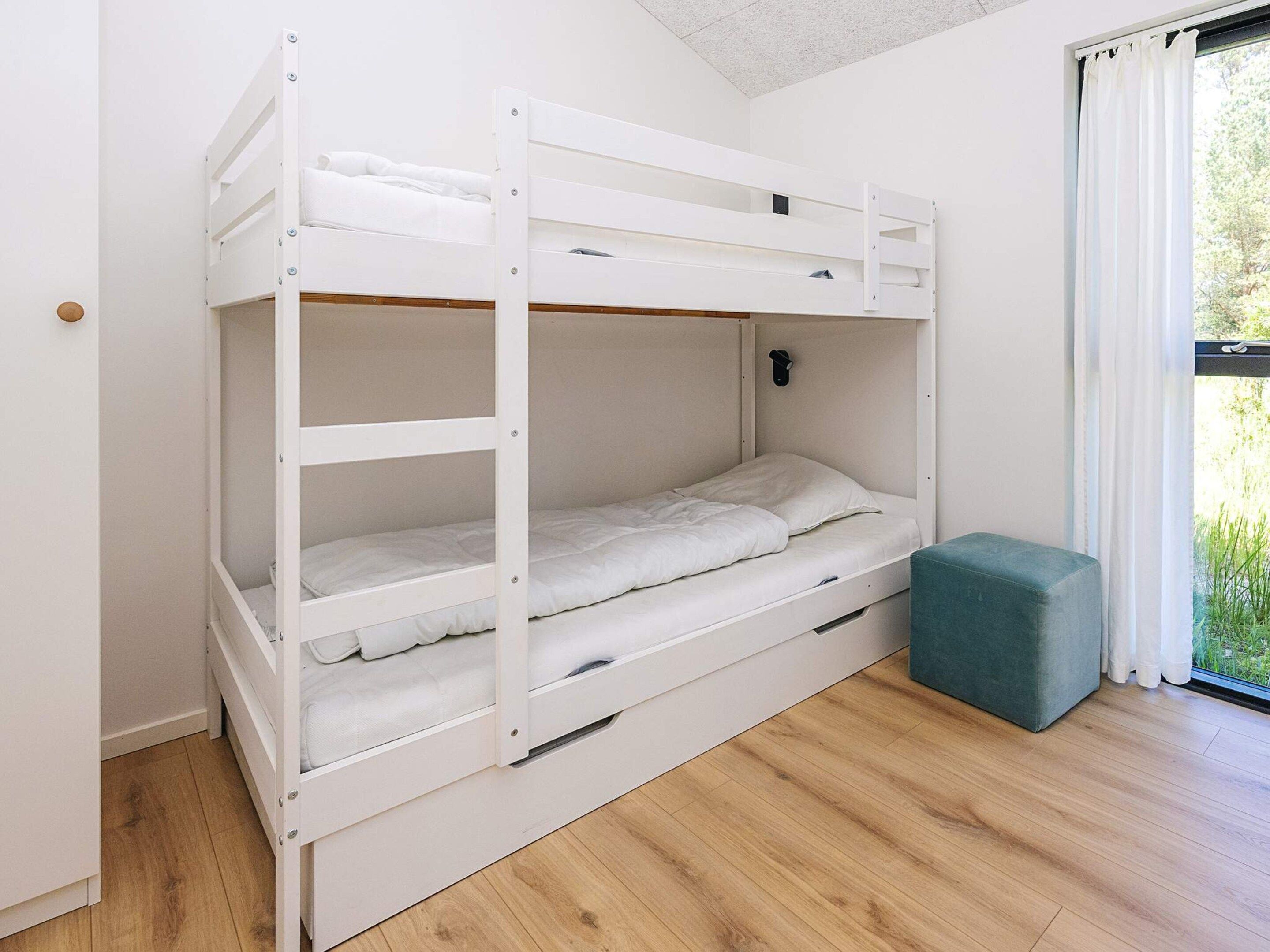 A bunk bed with a pull-out drawer, a small ottoman, and a window with a view of greenery.