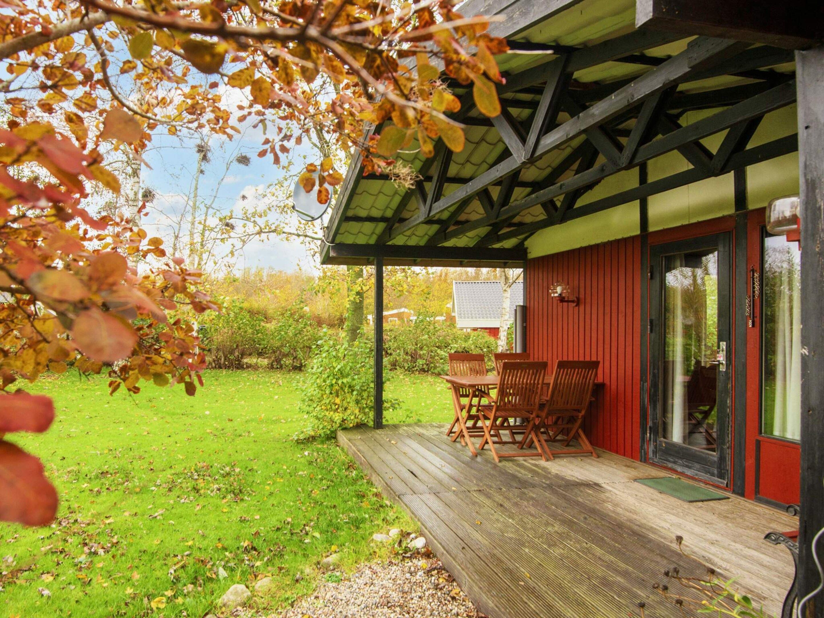 Ein überdachter Außenbereich mit Holzterrasse, ein rotes Gebäude mit Glasüren und ein Rasengrundstück.