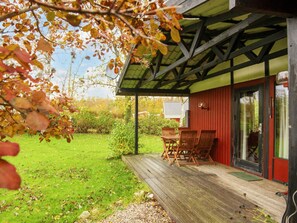 Un espace extérieur abrité avec une terrasse en bois, un bâtiment rouge doté de portes vitrées et une cour verdoyante.