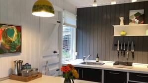 Een moderne keuken met een wit aanrechtblad, een zwarte achterwand en een hanglamp. Er is een raam met jaloezieën, een spoelbak en een plank met keukengerei.