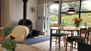 Een knus vertrek met een houtkachel, een eettafel met stoelen, en een groot raam met uitzicht op groen.