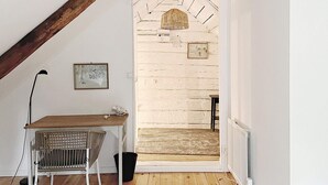 Una habitación con techo inclinado, un escritorio de madera, una silla, un radiador y una puerta que da a otra habitación.