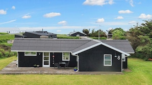 Une maison à deux étages, avec une façade noire et un toit gris, portant le numéro 8, dotée d’un petit patio et d’une porte donnant sur un porche abrité.