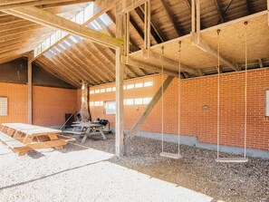 Een ruime binnenruimte met een hoge nok, zichtbare houten balken en een bakstenen muur. Er is een stenen open haard, een houten tafel en een schommel die aan het plafond hangt.