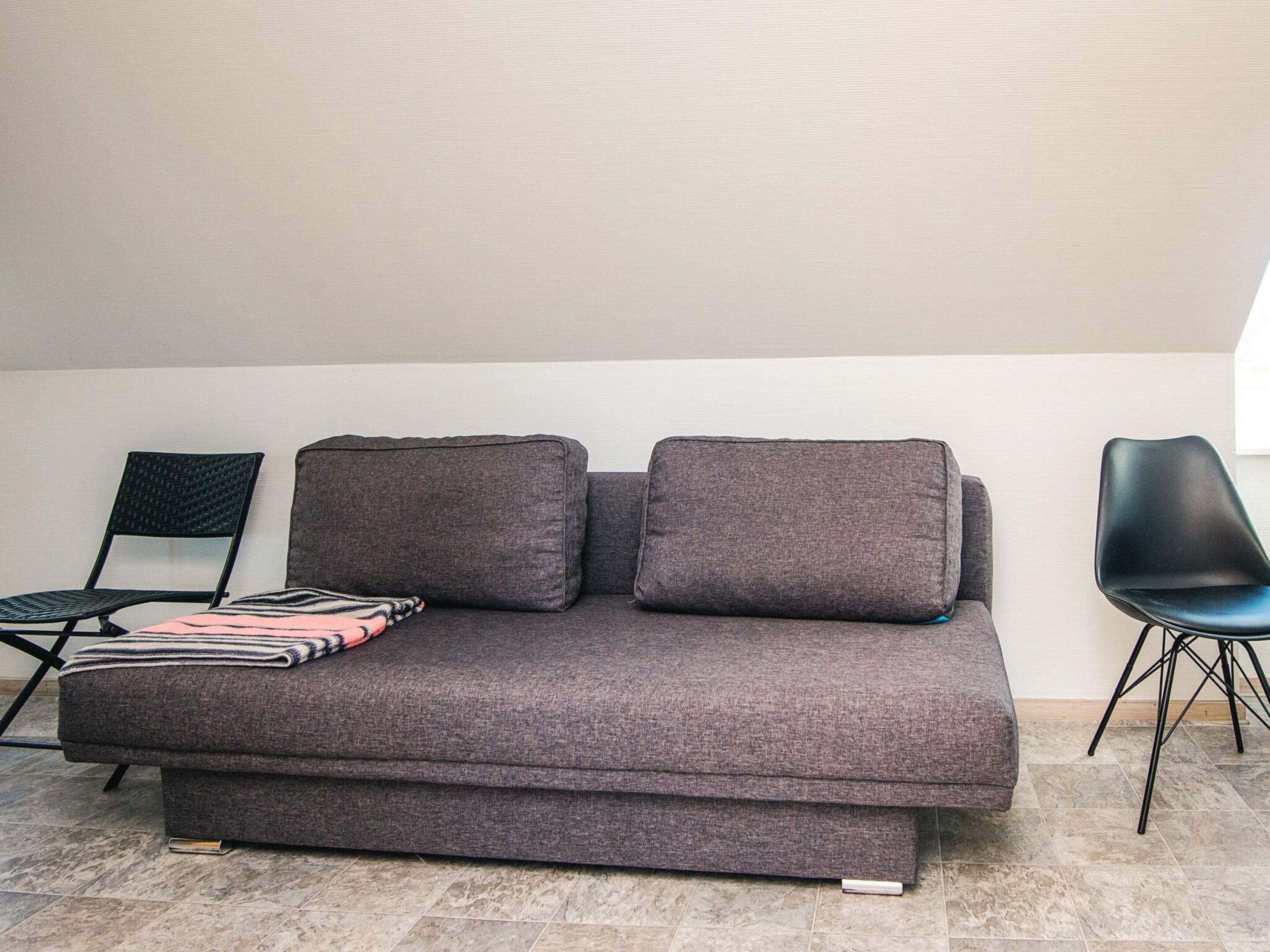 Ein graues Sofa mit zwei Kissen, einer gestreiften Decke und einem schwarzen Sessel in einem Raum mit Fliesenboden und schlichter Wand.