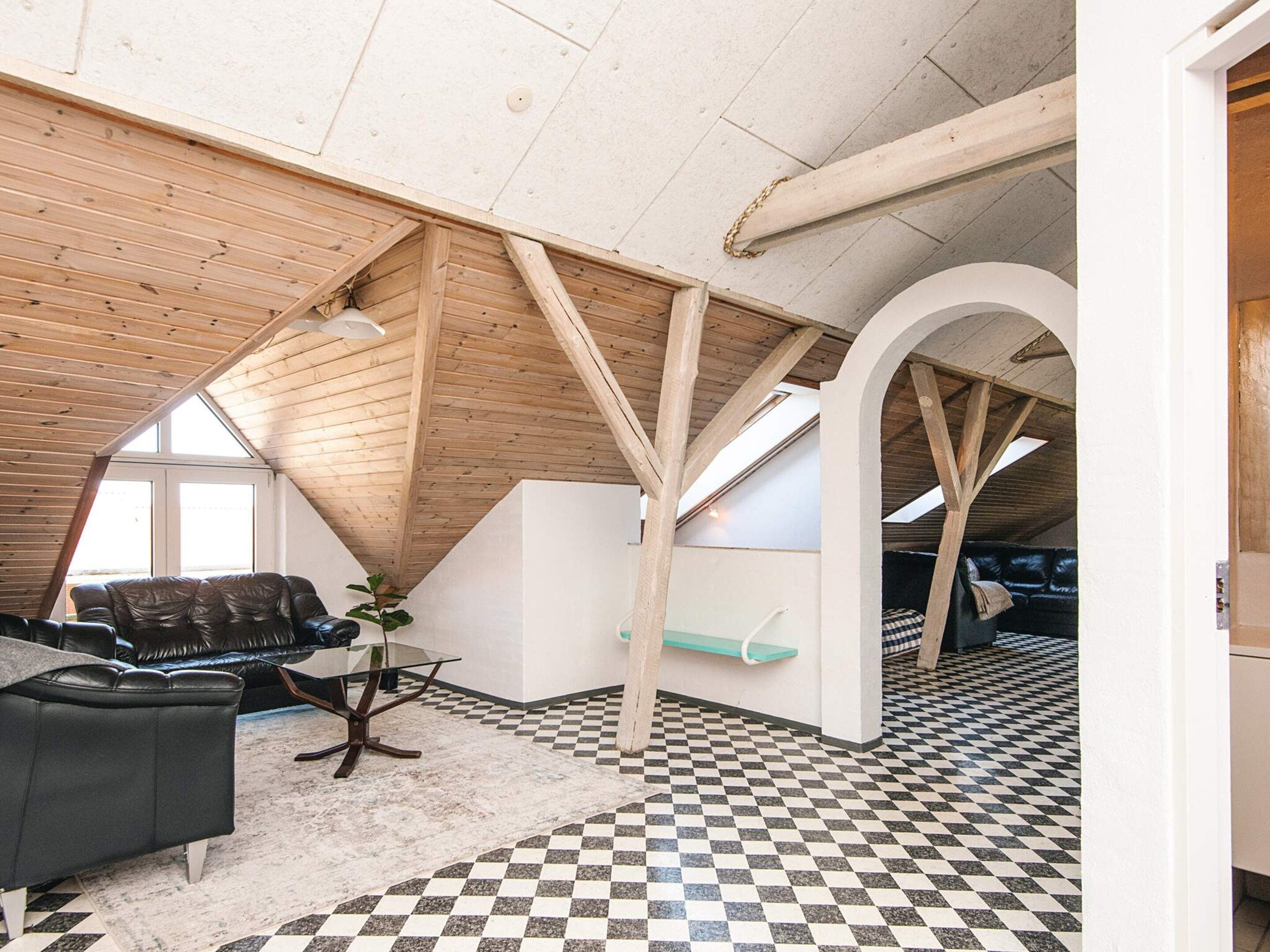 A loft-style room with a high ceiling, exposed wooden beams, a checkered floor, a leather sofa, and a glass table.