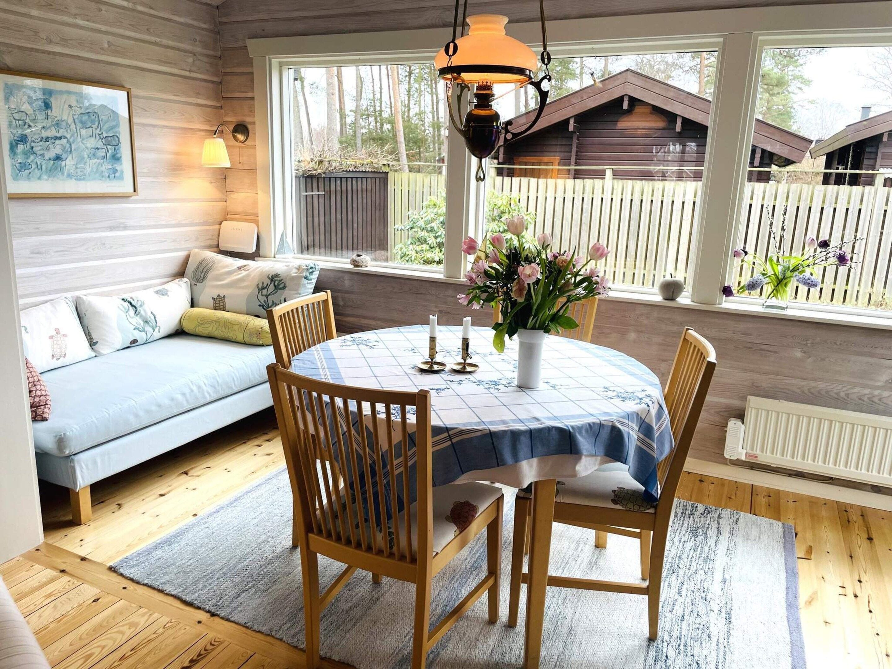 A cozy room with a round table, chairs, a sofa, and a window with a view of a wooden fence and trees.