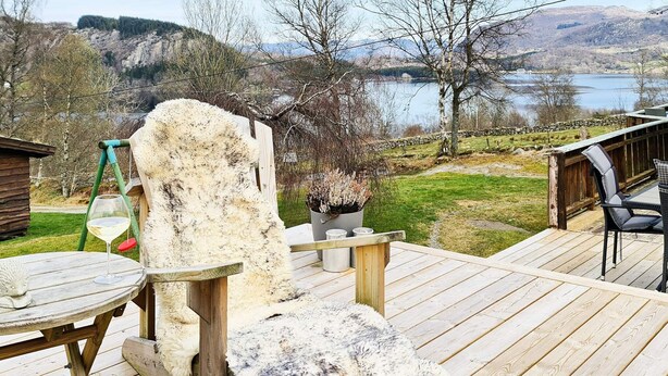 Une terrasse en bois avec une structure en pierre rustique, un verre de vin blanc et une vue sur un lac et des collines en arrière-plan.