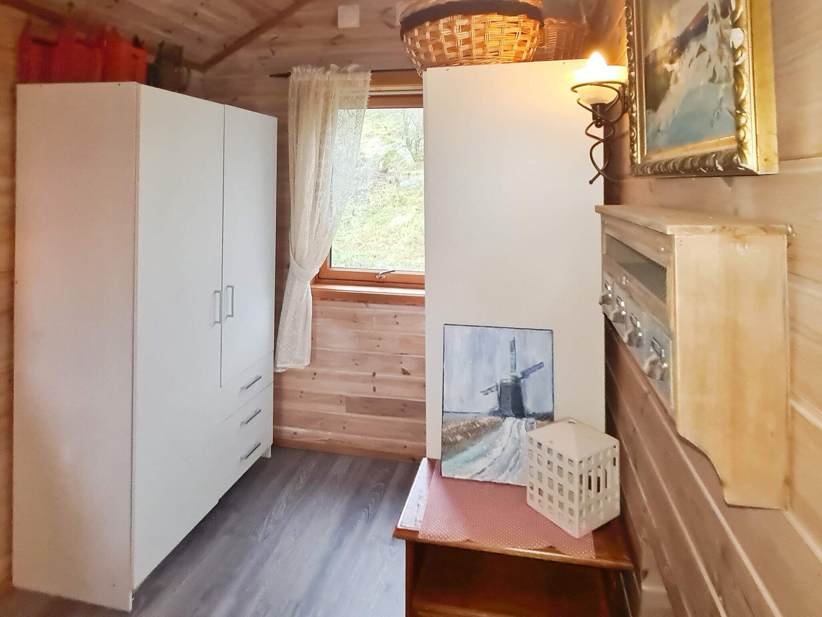 Una habitación con suelo de madera, un armario blanco, un ventanal con vistas a zonas verdes, un cuadro de un molino y una lámpara en la pared.