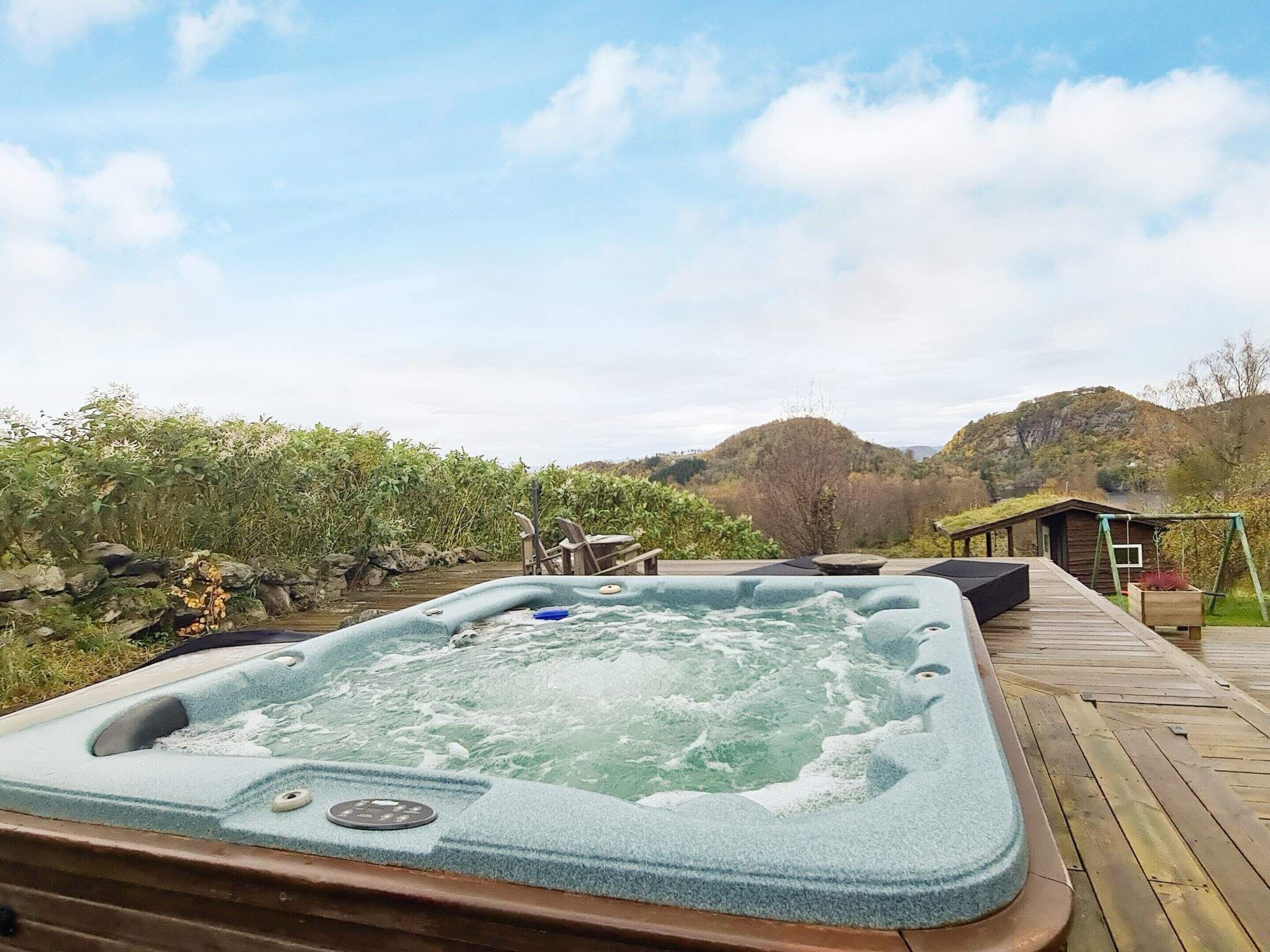 A hot tub on a wooden deck with a view of hills and a small cabin in the background.