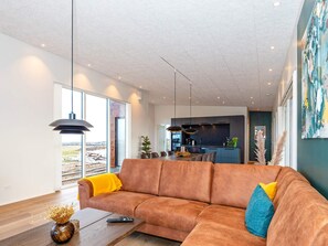 A modern living room with a large brown sofa, a wooden coffee table, and a view of the kitchen and landscape through large windows.