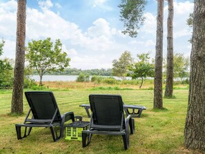 A grassy outdoor area with two black chairs and a small table, surrounded by trees and a view of a lake.