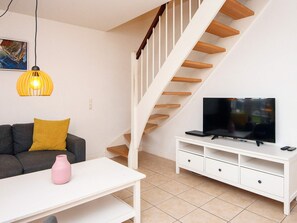 Una sala de estar con un sofá, un televisor sobre un mueble bajo, una escalera y una lámpara colgante.