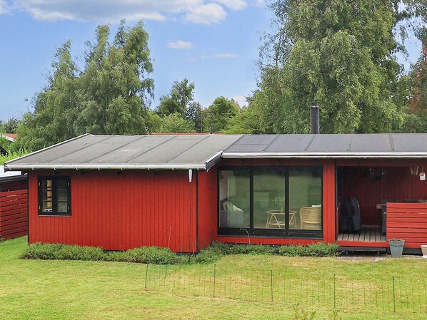 A red single-story building with a large glass door and windows, a flat roof, and a small porch area. - 4 Person Holiday Home in Vaeggerlose (Vaeggerlose)