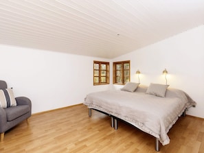 A bedroom with a bed, two bedside lamps, a gray armchair, and two windows with wooden frames.