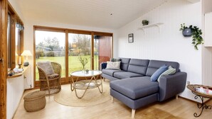 Un salon chaleureux avec un canapé d’angle gris, des fauteuils en osier, une petite table ronde et une grande porte-fenêtre coulissante donnant sur un jardin.