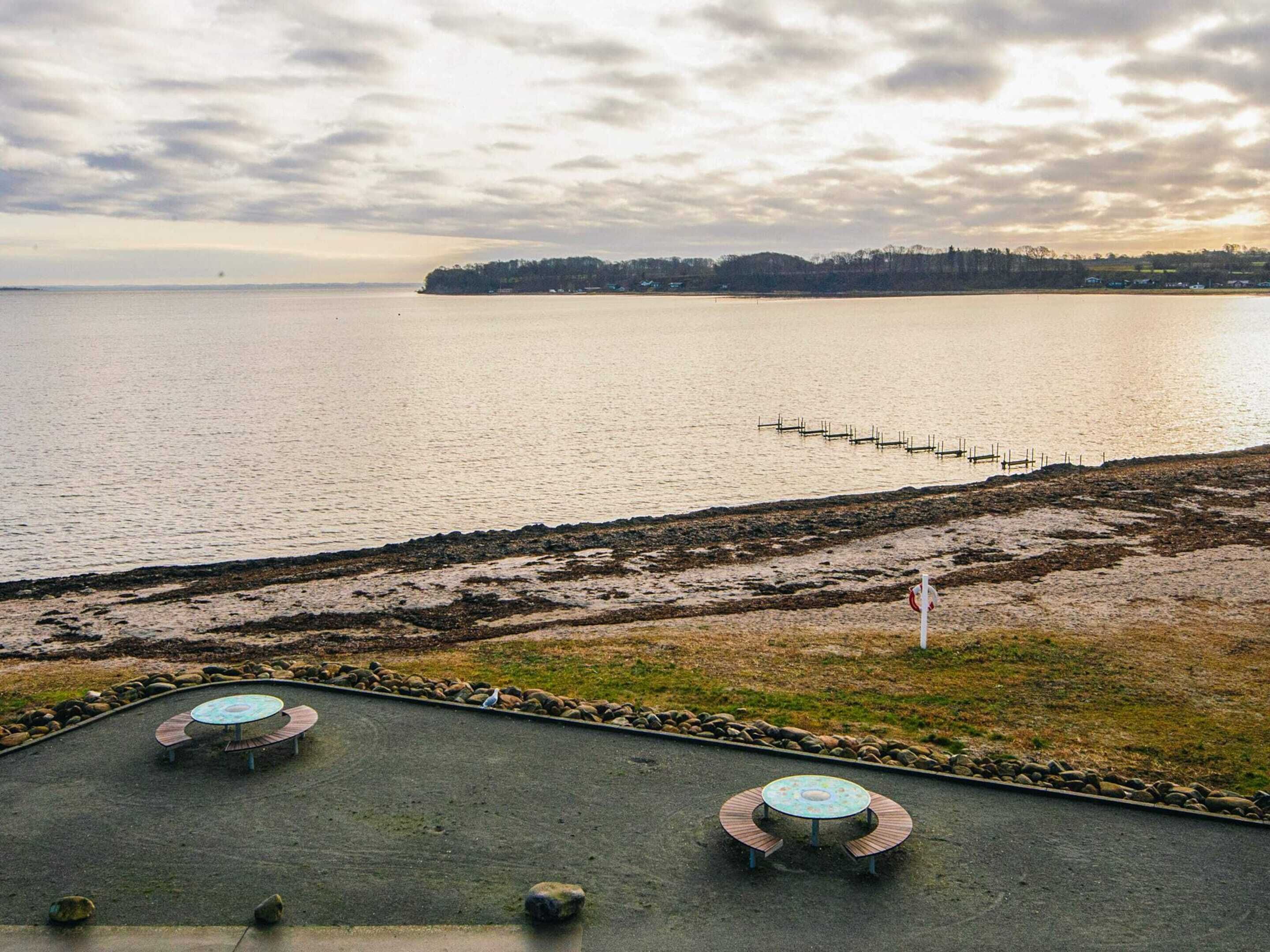 A waterfront view with a pier extending into the water, a grassy area with a circular seating arrangement, and a distant shoreline with trees.
