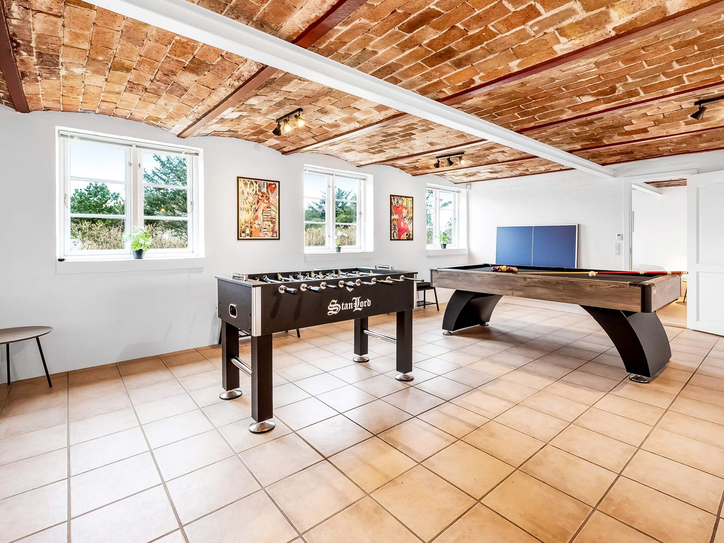 A room with a foosball table, pool table, and windows with views of greenery.