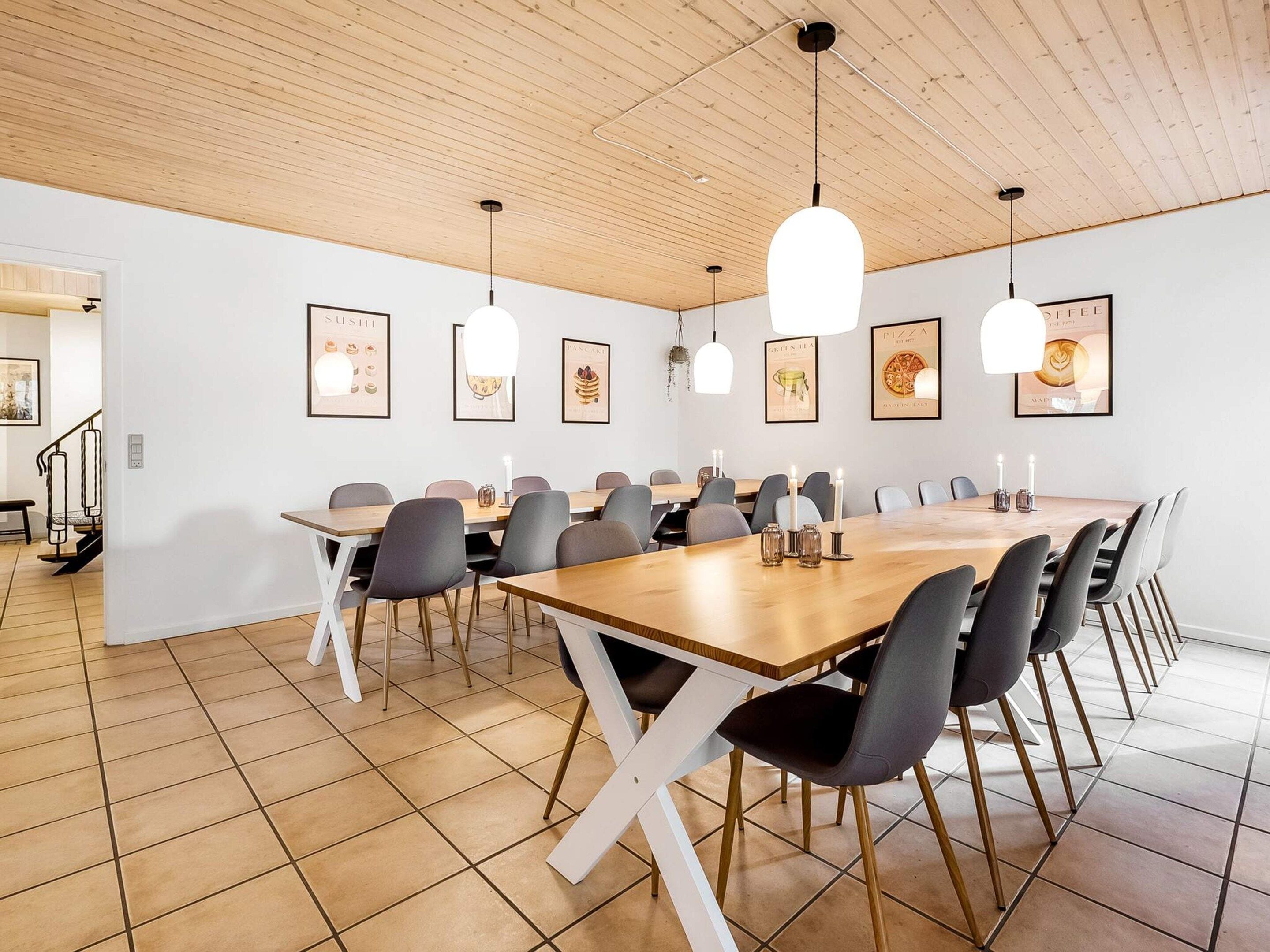 A dining area with a long wooden table, multiple chairs, pendant lights, and framed food posters on the wall.