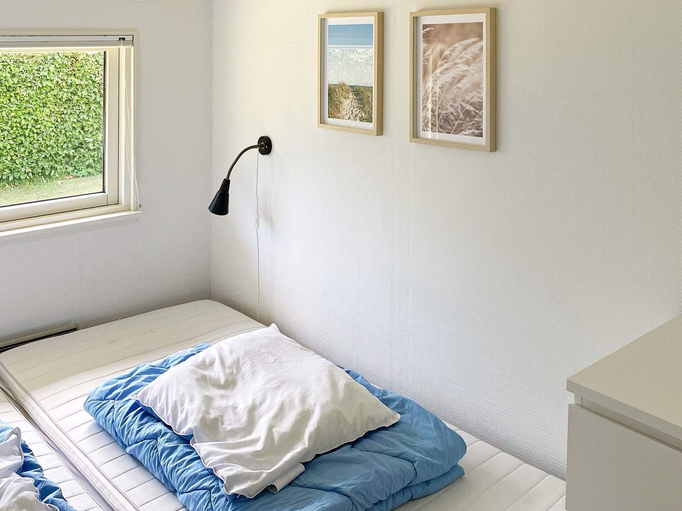 Una habitación pequeña con cama individual, ventana, lámpara de noche y dos cuadros en la pared.
