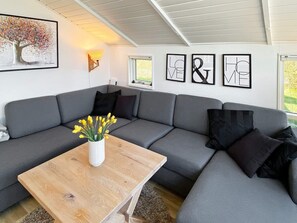 En moderne stue med en grå modulsofa, et træsofabord og en vase med gule blomster. Der er indrammede kunstværker på væggen, og et vindue med udsigt til grøn vegetation.