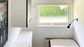 Ein gemütliches Zimmer mit Bett, Schreibtisch, Fenster mit Blick auf Grünflächen und einer Wandlampe.