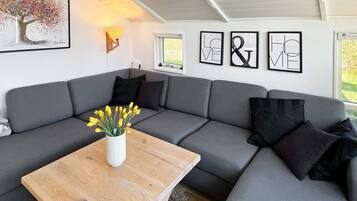 A modern living room with a grey sectional sofa, a wooden coffee table with a vase of yellow flowers, and wall art including a tree painting and framed text.