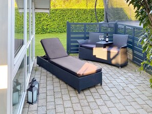 Een terras met een lounge stoel en een bank, een kleine tafel met een fles en glazen, en een lantaarn.