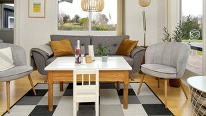 A modern living room with a grey sofa set, a wooden dining table, and a large window offering a view of the outdoors. - 6 Person Holiday Home in Olsted (Frederiksvaerk)