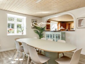A dining area with a long table and white chairs, a kitchen with wooden cabinets, and a window with flowers. - 4 Star Holiday Home in Vaeggerlose (Vaeggerlose)