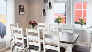 Un coin repas avec une table en bois et des chaises blanches, un vase avec des fleurs rouges et une fenêtre dont l’extérieur est rouge.