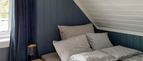 A bedroom with a sloped ceiling, a bed with pillows, a bedside table with a lamp, and a window with curtains.