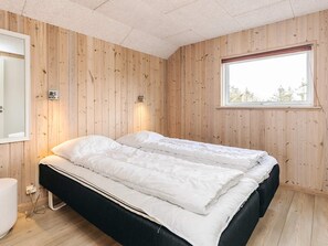 A room with wooden panel walls, a bed with white bedding, a window with a view, and a wall-mounted light fixture.
