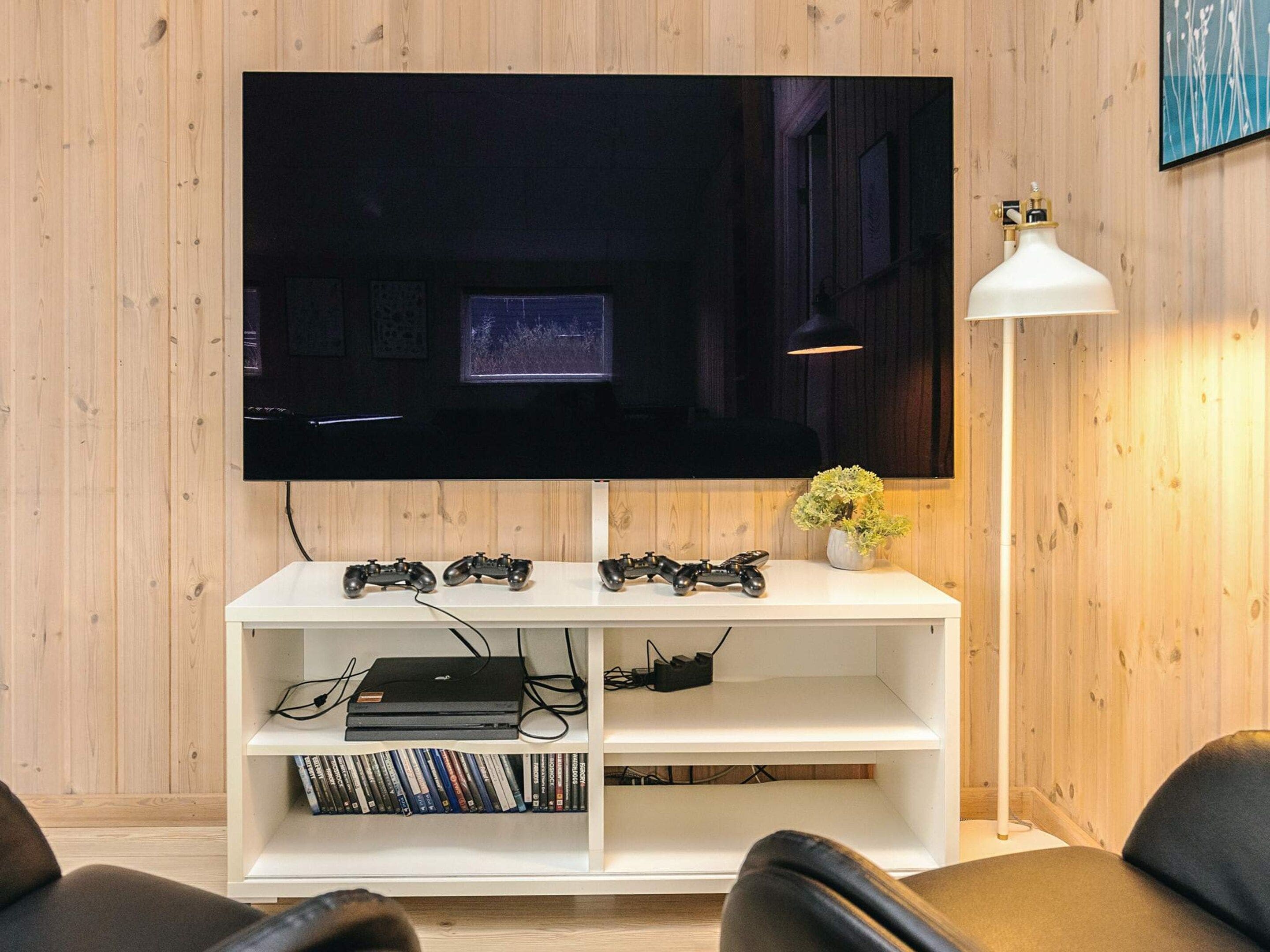 A living room with a flat-screen TV mounted on a wooden wall, a white entertainment unit with gaming consoles and a laptop, a floor lamp, and a potted plant.