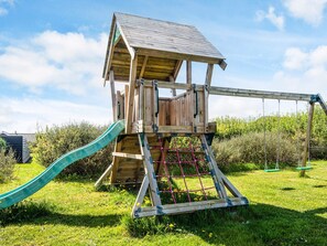 A wooden playhouse with a slide and a swing set in a grassy area.