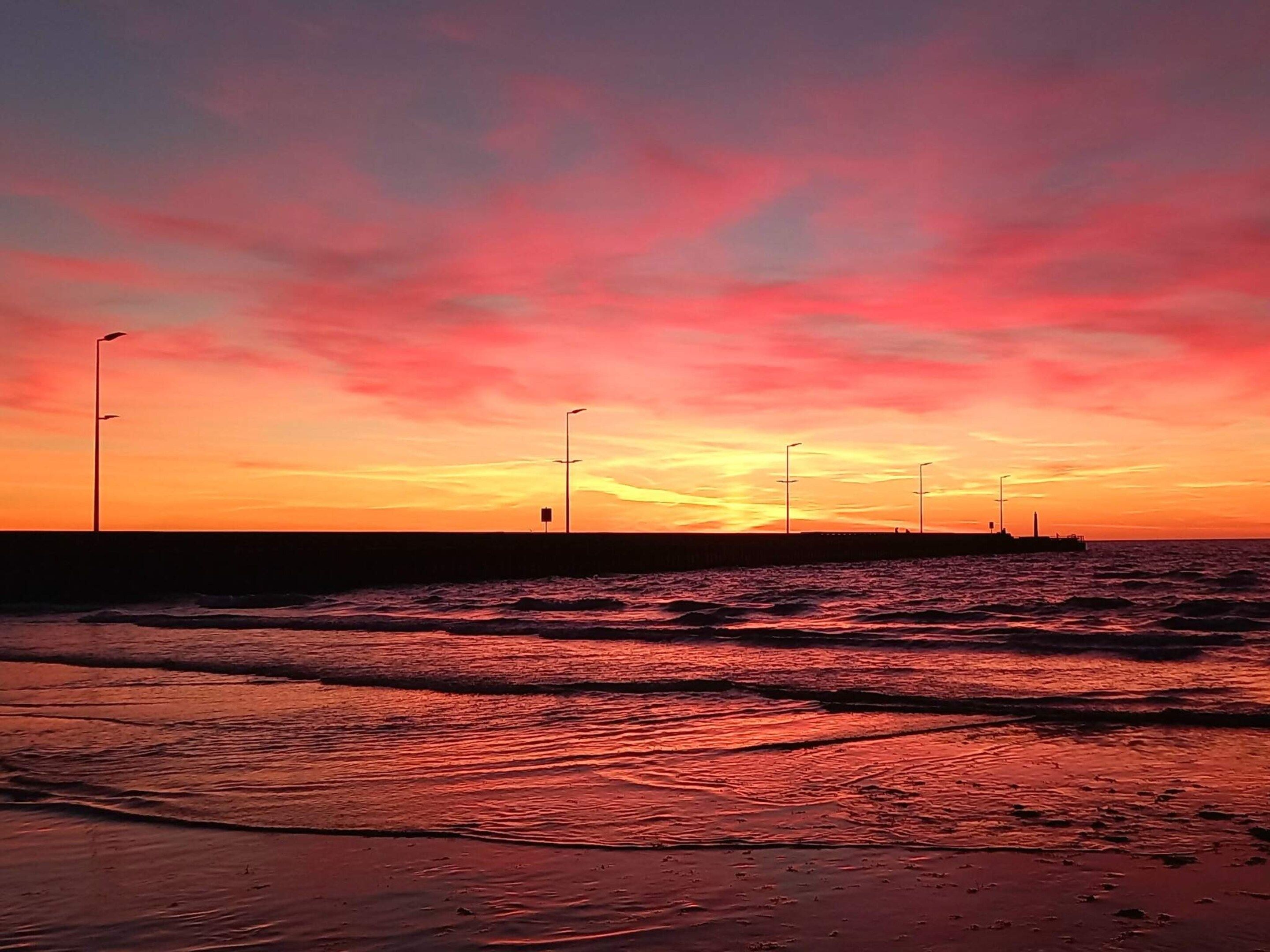 Ein Sonnenuntergang über einem Strand mit Wellen, einem Steg und Straßenlaternen.